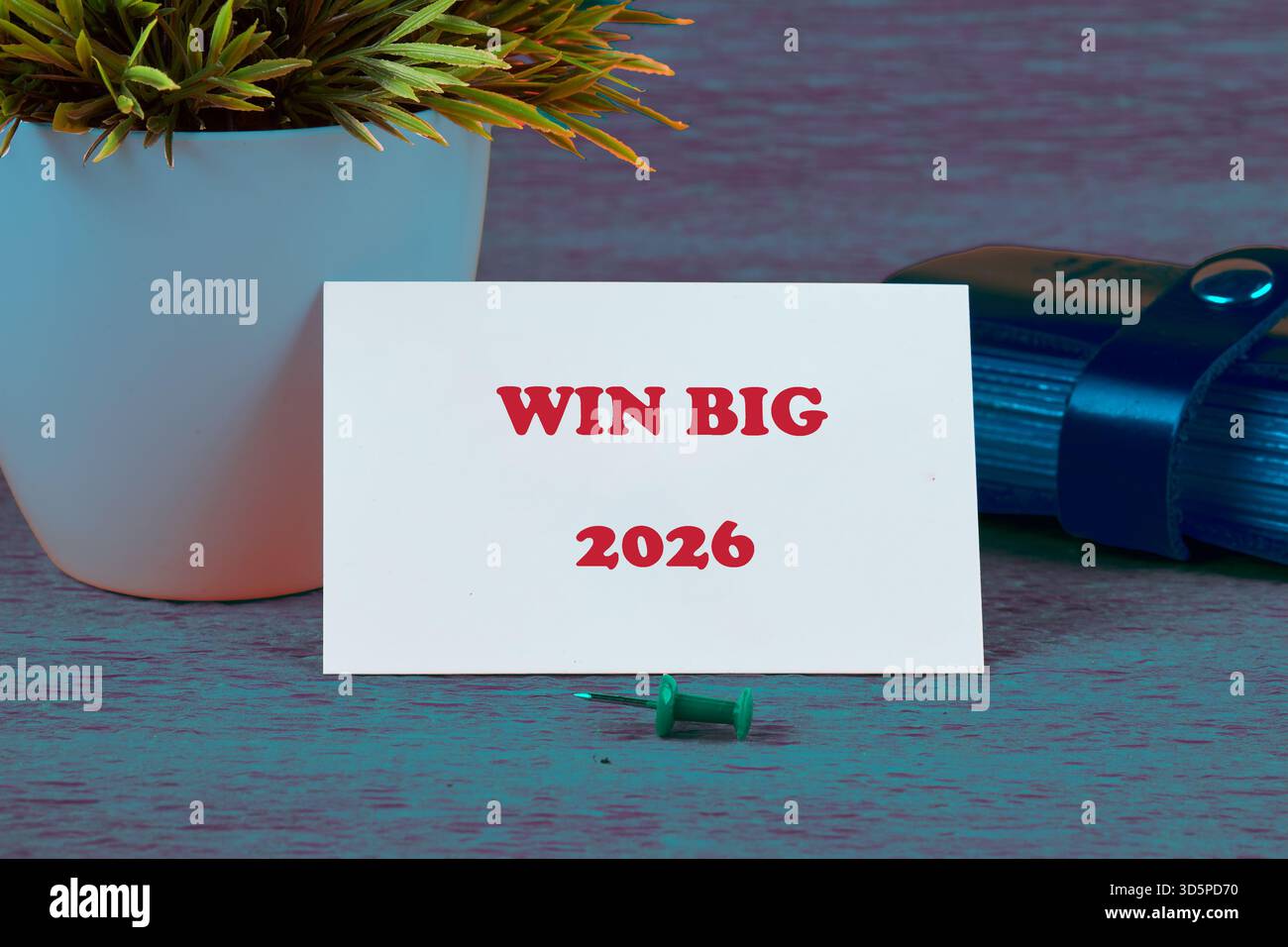 Eine weiße Karte zeigt Win Big 2026 in rotem Text an. Daneben sitzt eine kleine Pflanze in einem weißen Topf. Eine blaue Rolle wird mit einem Band an der Seite mit einem t gesichert Stockfoto