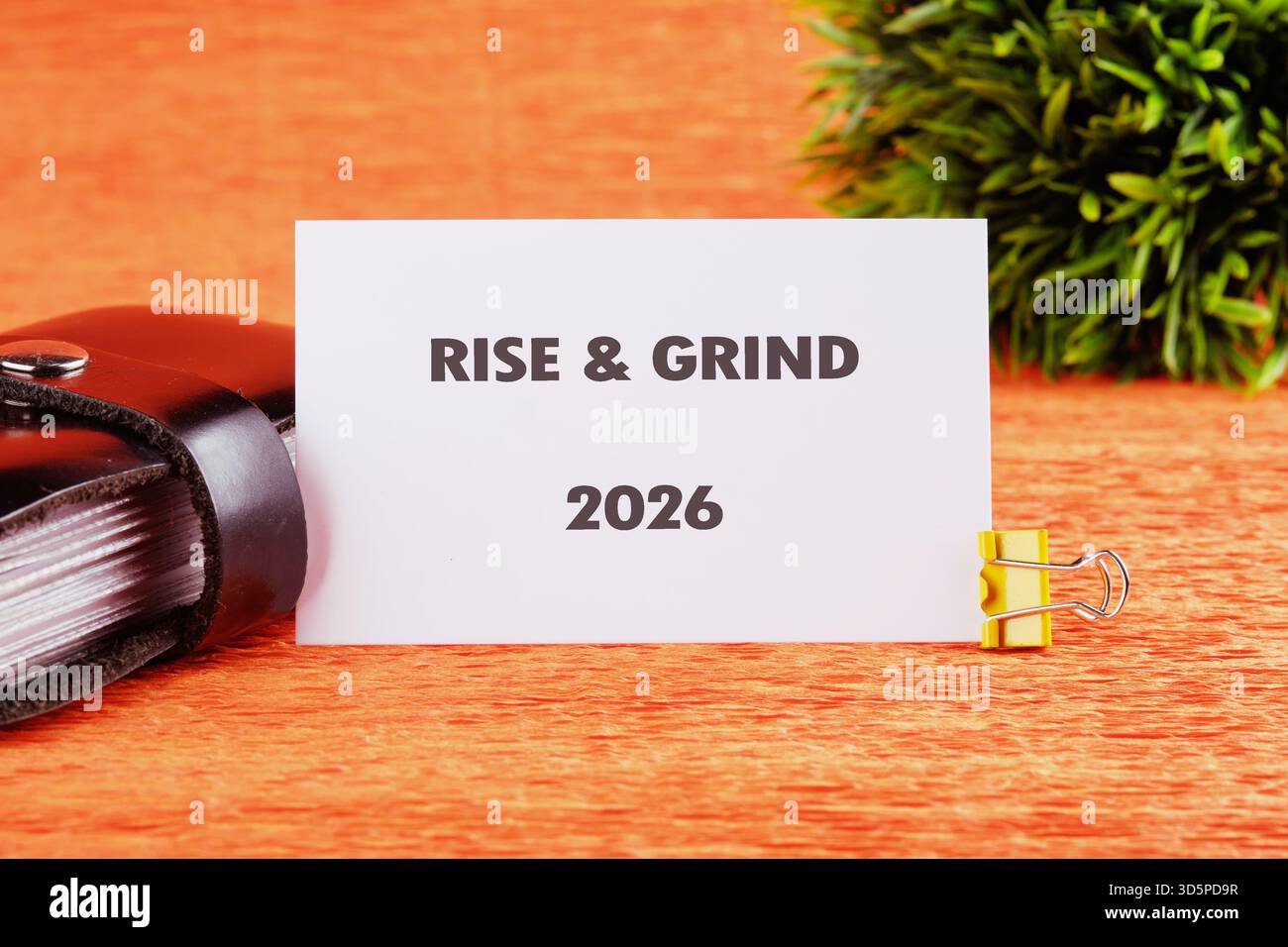 Eine Karte mit der Phrase RISE GRIND 2026 wird mit einem goldenen Binderclip geclippt. Es sitzt auf einer strukturierten orangefarbenen Oberfläche, neben einem schwarzen Buch und Grün Stockfoto