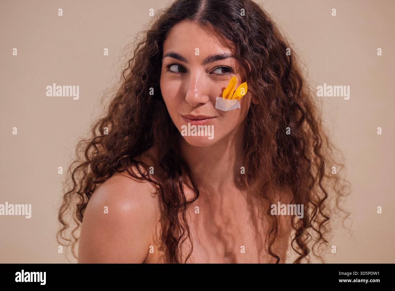 Die junge Frau mit lockigem Haar posiert spielerisch mit buntem Papier auf der Wange und hebt Kreativität und künstlerischen Ausdruck in minimalistischer Atmosphäre hervor Stockfoto