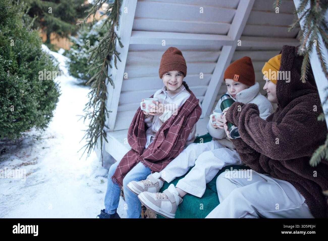 Kinder sitzen in einem charmanten Outdoor-Schutzhaus und schlürfen warme Getränke, umgeben von schneebedeckten Bäumen, die ein Gefühl von Freude und Wärme während der Zeit wecken Stockfoto