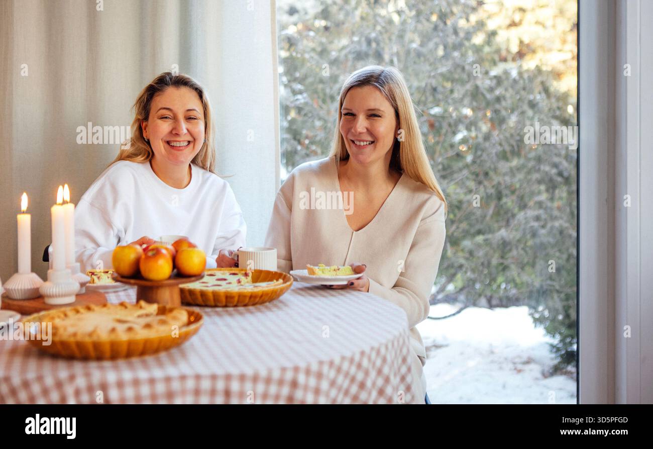 Zwei fröhliche Frauen sitzen an einem Tisch, der mit Gebäck und Äpfeln geschmückt ist, und genießen warme Drinks in einer gemütlichen Winterumgebung, die Glück und Wärme ausstrahlt Stockfoto