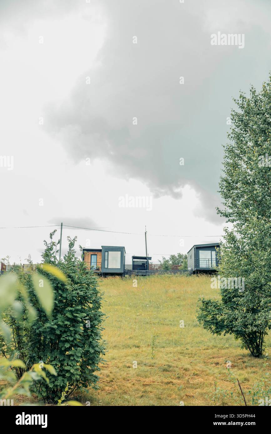 Moderne modulare Häuser mit großen Fenstern, auf grünem Hügel, Gras, Büsche unter bewölktem Himmel, zeitgenössische Architektur für Öko-Wohnen, Landschaft Stockfoto