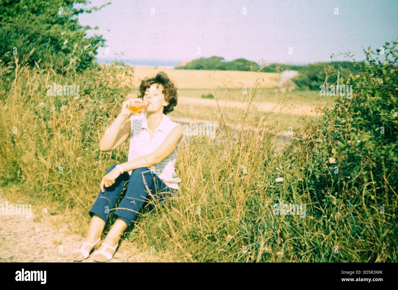 Eine Frau sitzt auf dem Gras am Wegesrand und stillt ihren Durst. [Automatisierte Übersetzung] Stockfoto