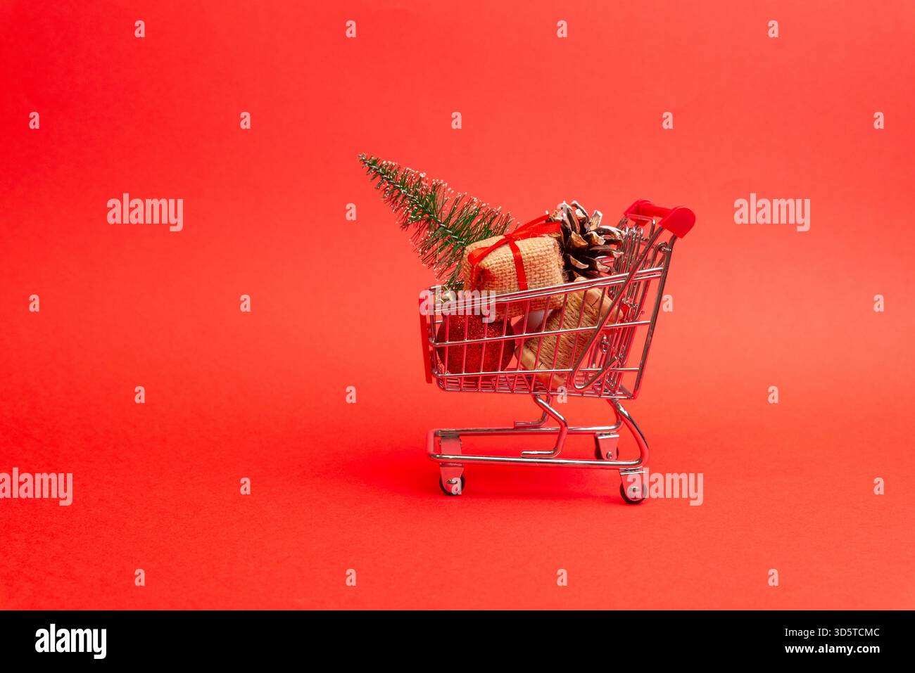 Mini-Einkaufswagen mit Weihnachtsgeschenken, Tannenzapfen und Dekorationen auf einem sauberen roten Hintergrund für Weihnachtsthemen Stockfoto