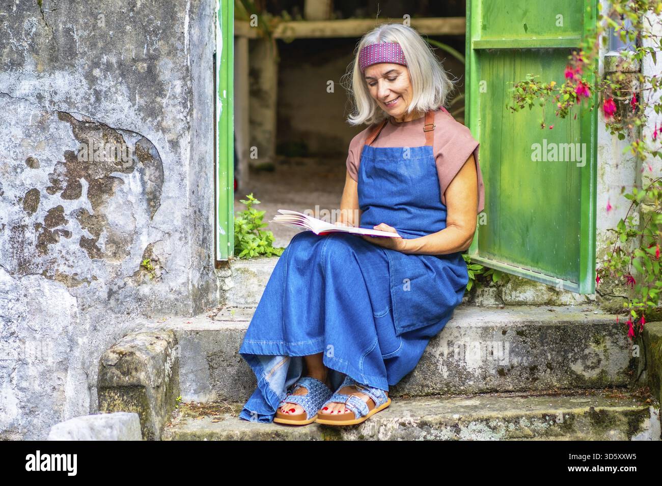 Eine Seniorin aus Denim sitzt auf alten Steintreppen in einem sonnigen Garten, liest friedlich ein Buch, lächelt und genießt einen ruhigen Sommermoment von lei Stockfoto