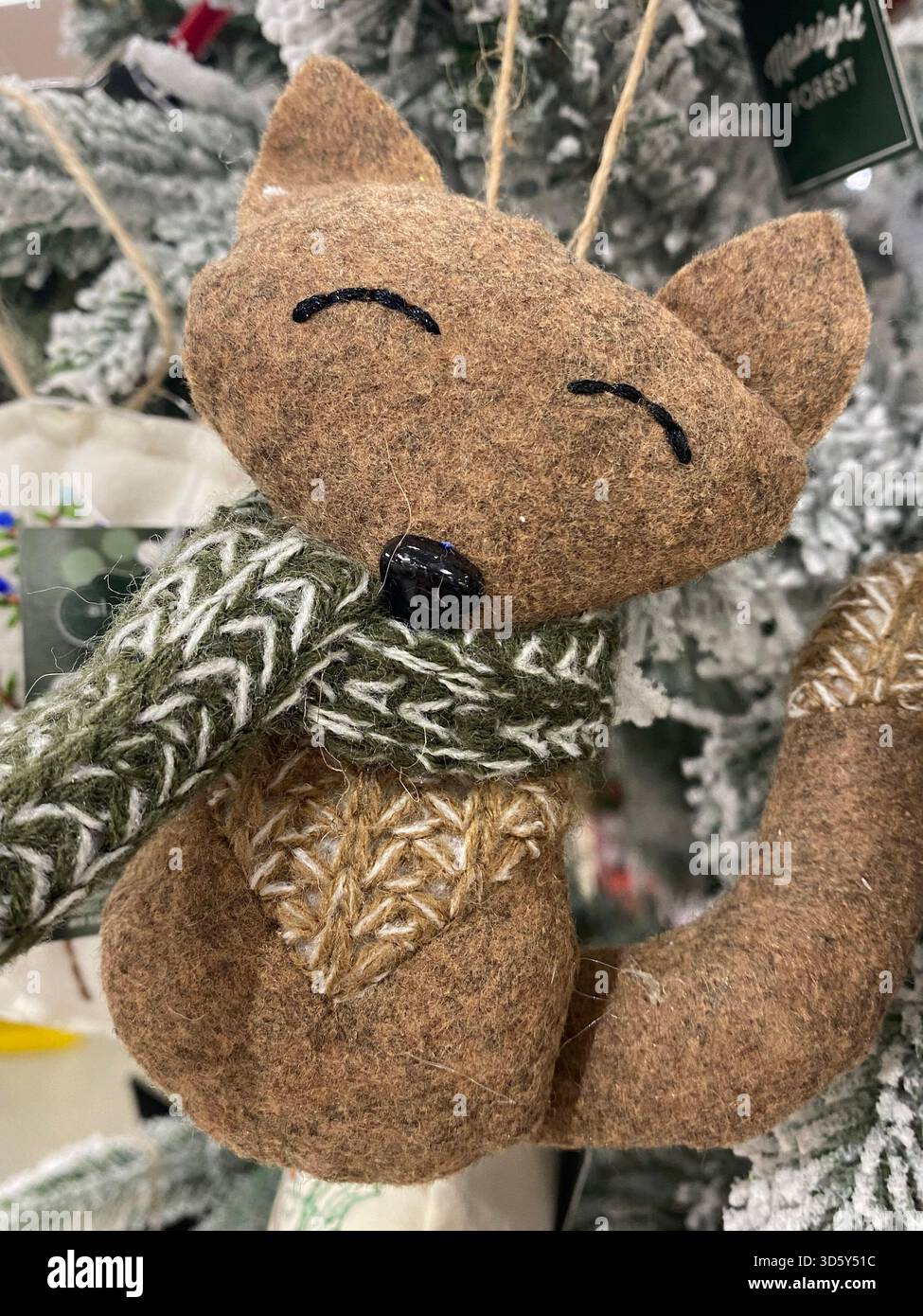 Ein weicher Fuchsschmuck aus Filz mit gestickten Details, ein grüner Strickschal hängt an einem schneebedeckten Weihnachtsbaum. Handgefertigte Textur, Einrichtung im Winter. - Smartphone-aufgenommenes Stockfoto