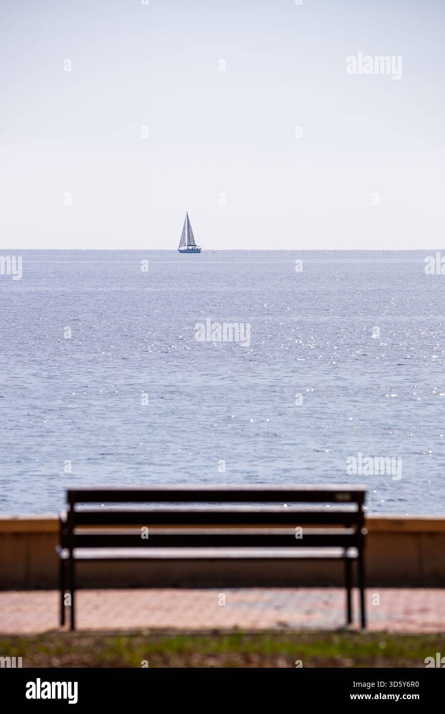 Eine ruhige, sonnendurchflutete Szene mit einem kleinen Segelboot, das über den glitzernden Horizont des Mittelmeers gleitet, hinter einer verschwommenen Holzbank Stockfoto