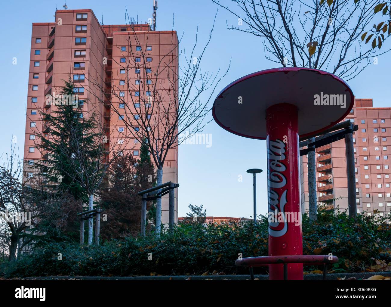 Einsamer Coca-Cola-Stehtisch vor DDR-Plattenbau-Apartmentblöcken im Berliner Ernst-Thälmann-Park bei Sonnenuntergang im Spätherbst. Stockfoto