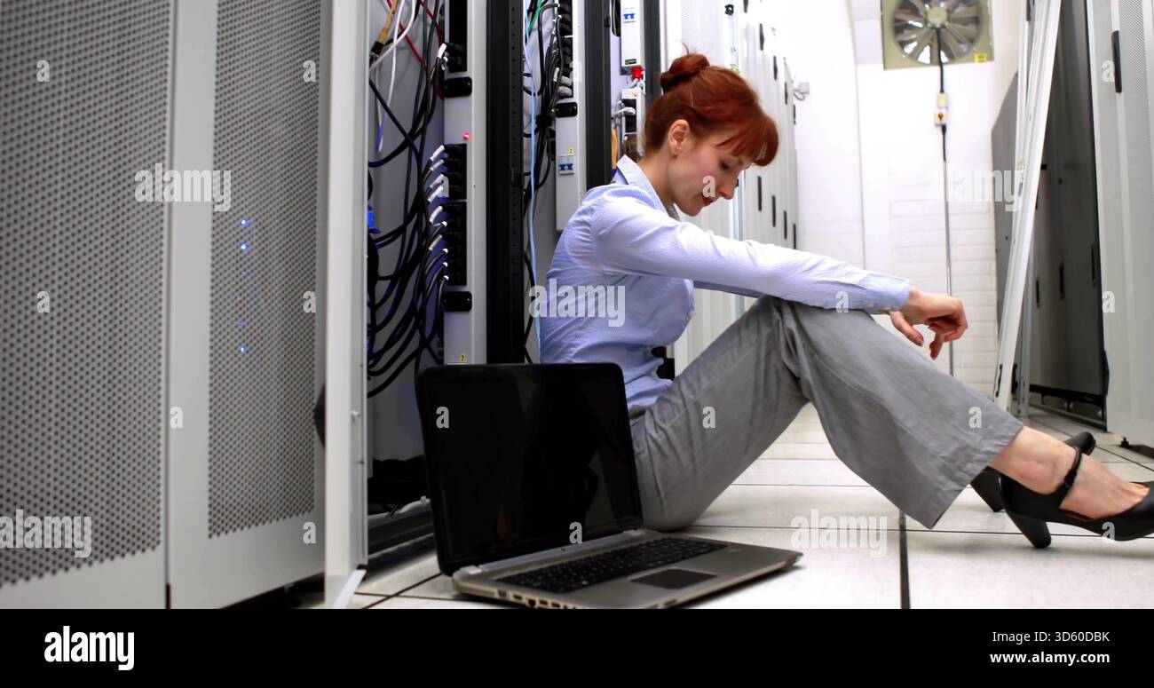 Sitzende Frau in blauem Hemd, die mit Kabeln zwischen Serverschränken im Rechenzentrum einen Laptop überprüft Stockfoto