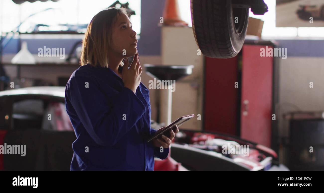 Untersuchung der Frau in blauen Overalls mit Tablettenhalter und Beurteilung des hängenden Rades im Garagenplatz. Mechaniker, Werkstatt, Lift, Auto, Werkzeuge, Gerät, Diagnose Stockfoto