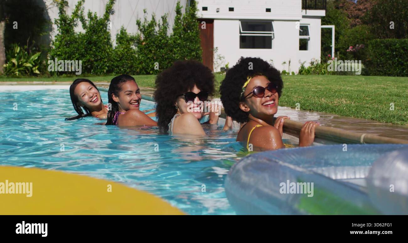 Vier Frauen in Badeanzügen, die im Pool im Hinterhof lächeln, mit gelbem Schwimmer Stockfoto