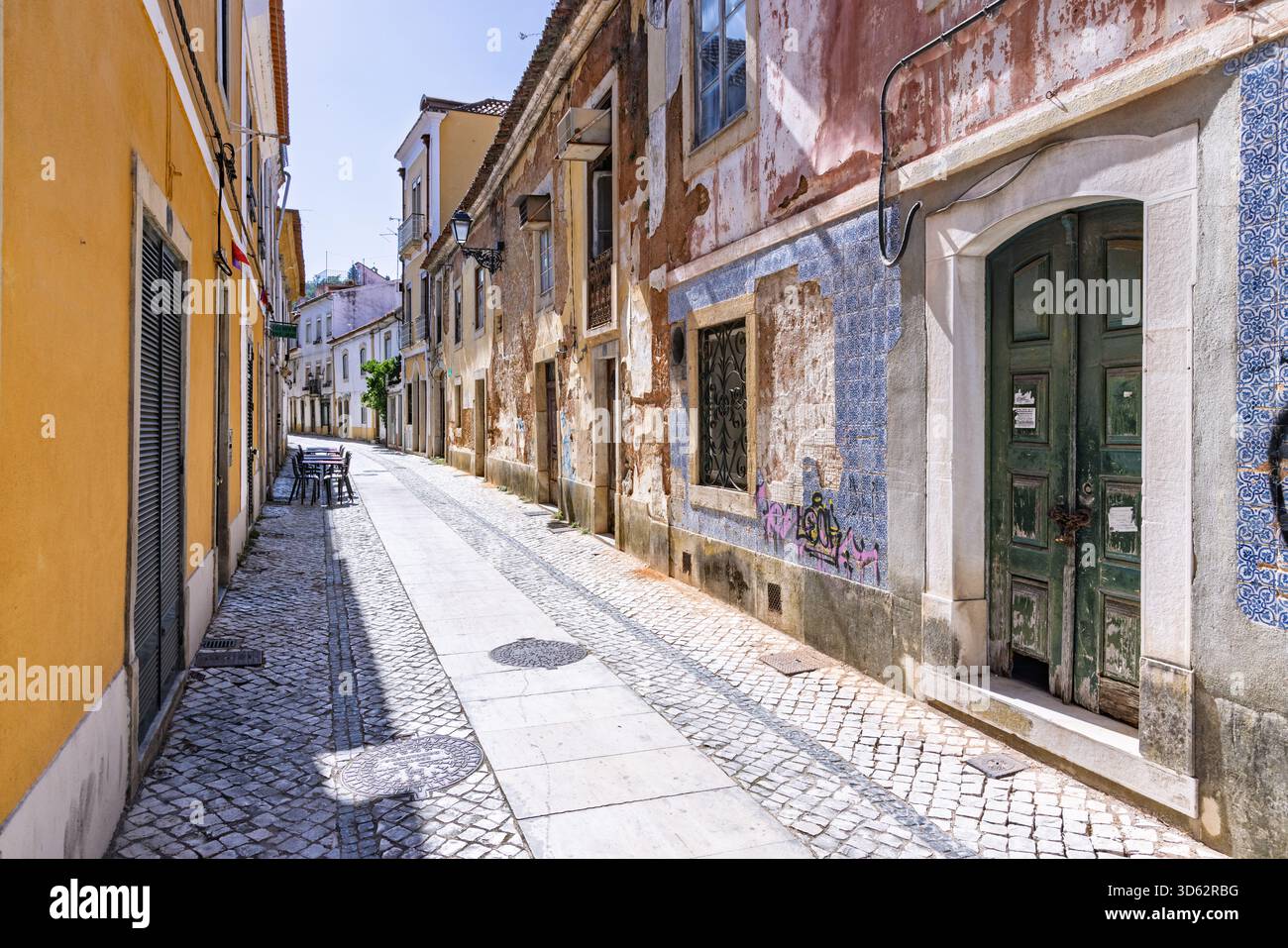 Eine bezaubernde, sonnendurchflutete Kopfsteinpflasterallee im historischen Zentrum von Tomar, Portugal, mit alten Gebäuden mit verblassten Fliesen und einer grünen Holztür. Stockfoto