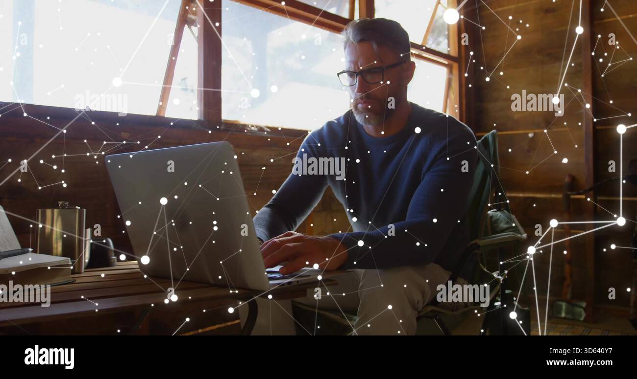 Arbeitender Mann mit marineblauem Pullover und Brille, der am Laptop am Kabinentisch schreibt, mit Reisebecher Stockfoto