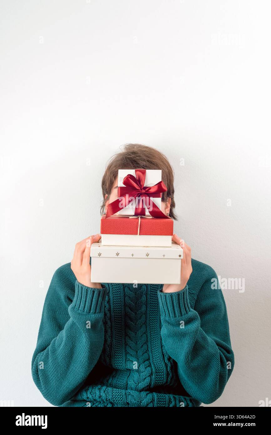 Mädchen in einem blauen Strickpullover, der einen hohen Stapel von rot-weißen Geschenkkartons mit Schleifen hält und ihr Gesicht vollständig vor einer weißen Wand bedeckt. Stockfoto