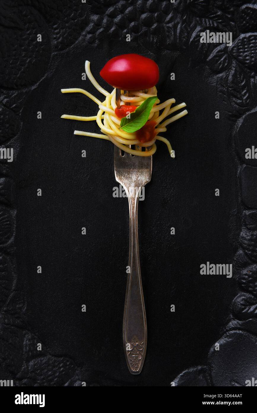 Eine Gabel mit Nudeln, Tomatensauce, einem Basilikumblatt und einer Kirschtomate auf einer schwarzen Servierplatte. Stockfoto