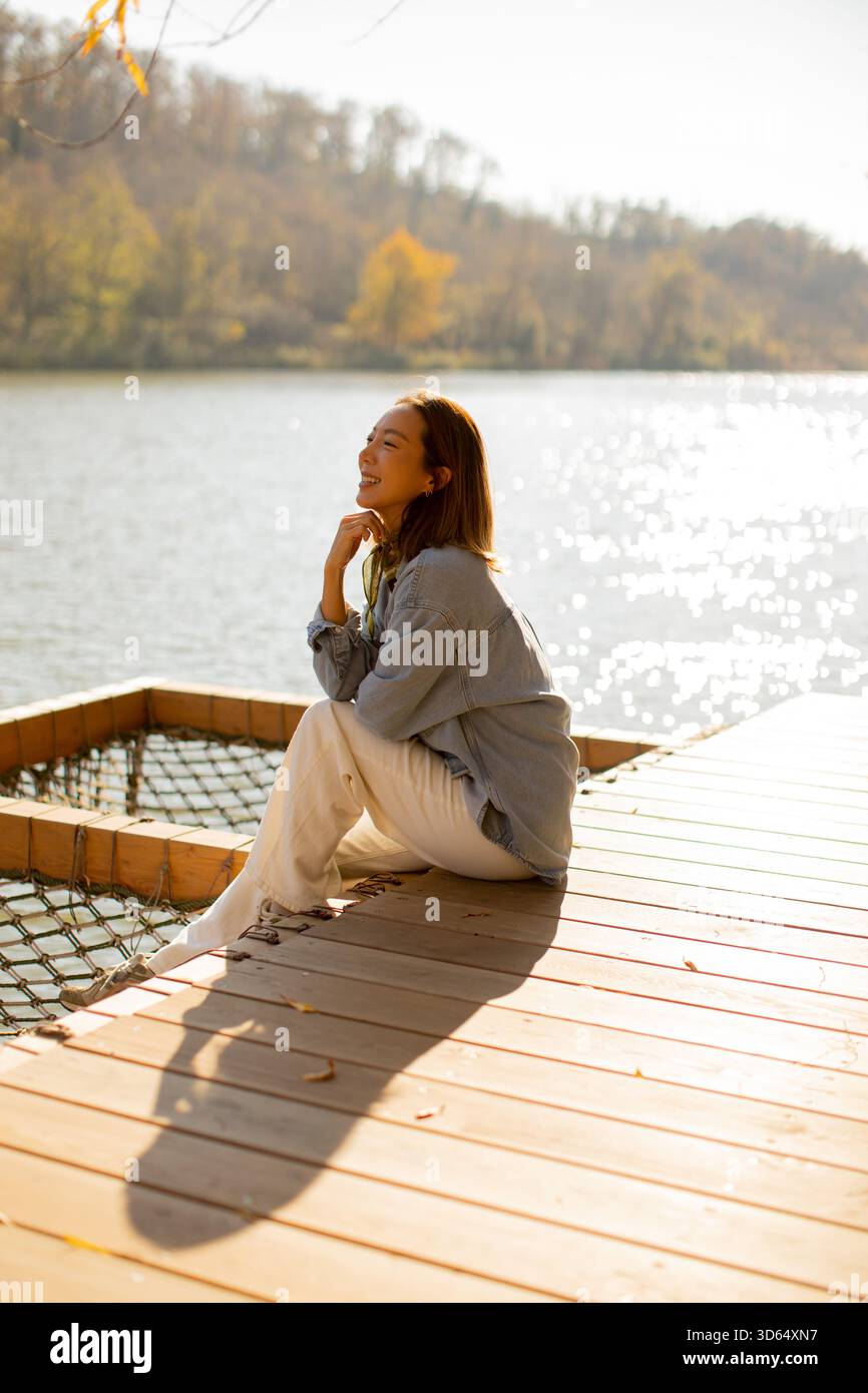 Eine junge Frau entspannt sich auf einem hölzernen Dock an einem ruhigen See, in der warmen Nachmittagssonne, umgeben von der Natur. Stockfoto