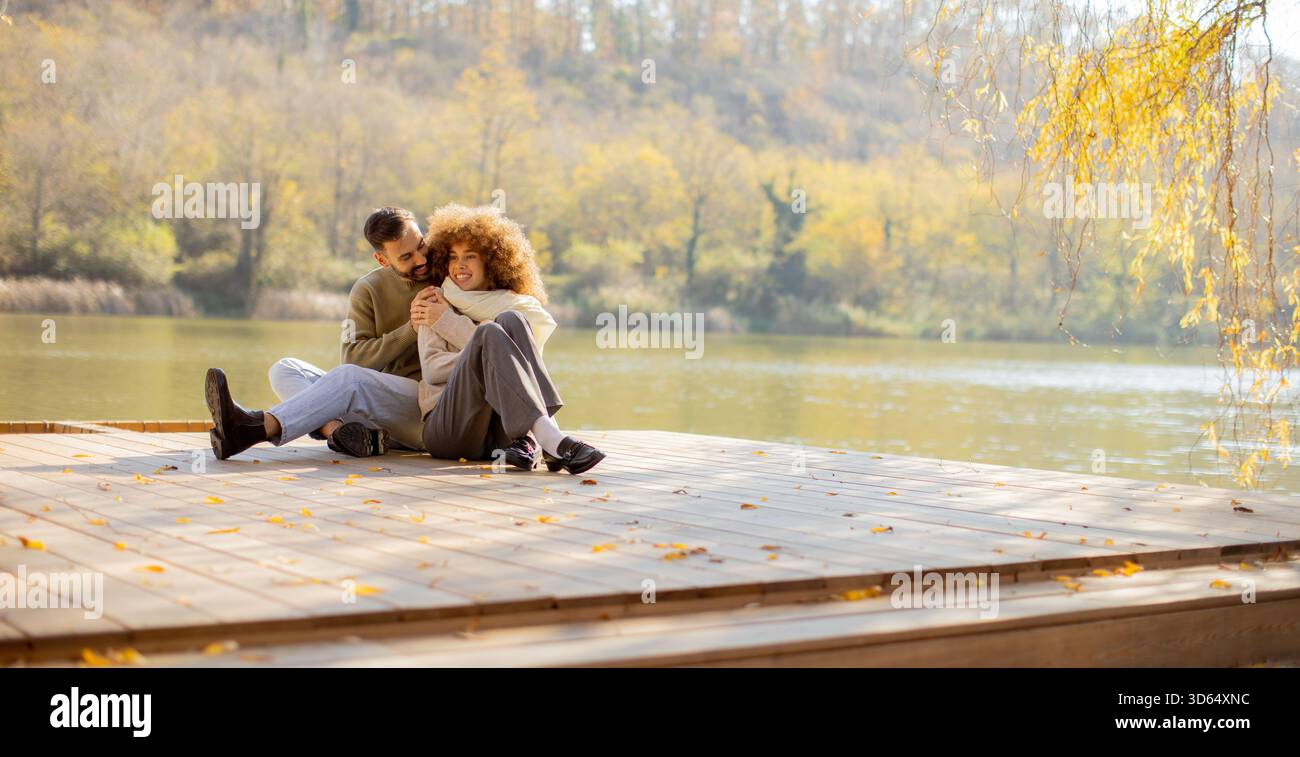 Ein junges Paar sitzt auf einem hölzernen Dock an einem ruhigen See. Lebhafte Herbstblätter umgeben sie, während sie intime Momente spielerisch umarmen und teilen. Stockfoto