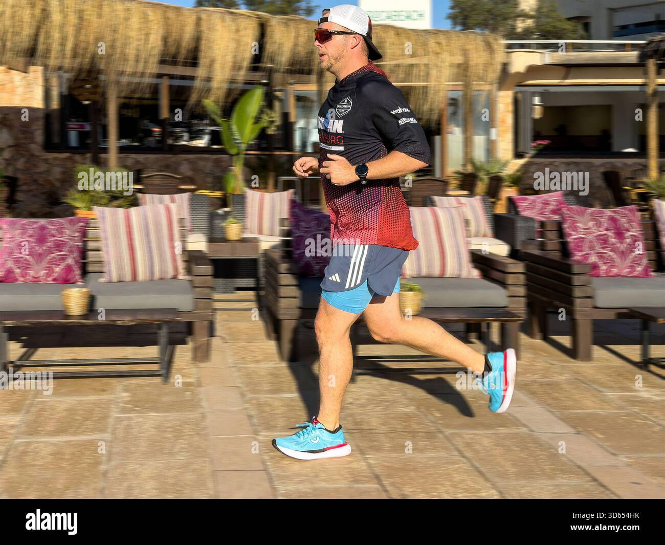 Mann in sportlicher Kleidung, der an einem sonnigen Tag an der Can pastilla mallorca spanien vorbei an einem Lounge-Bereich im Freien mit gepolsterten Sitzgelegenheiten und tropischem Dekor joggt Stockfoto