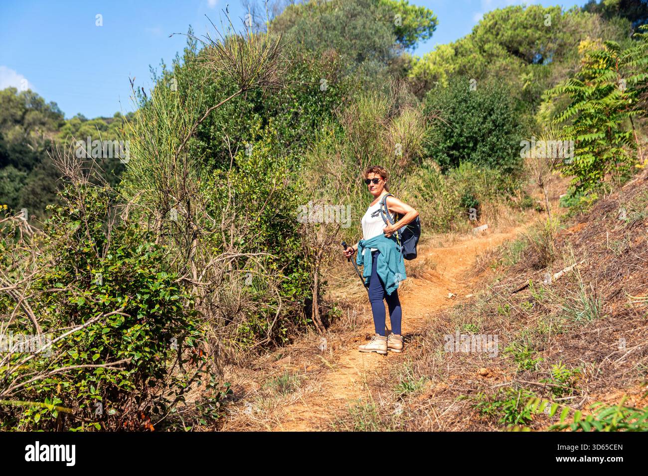 Seniorin, die auf einem gewundenen, unbefestigten Pfad steht, umgeben von üppig grünen Bäumen und Bürste, trägt einen Rucksack, eine Sonnenbrille und Aktivkleidung und genießt einen Auftritt Stockfoto
