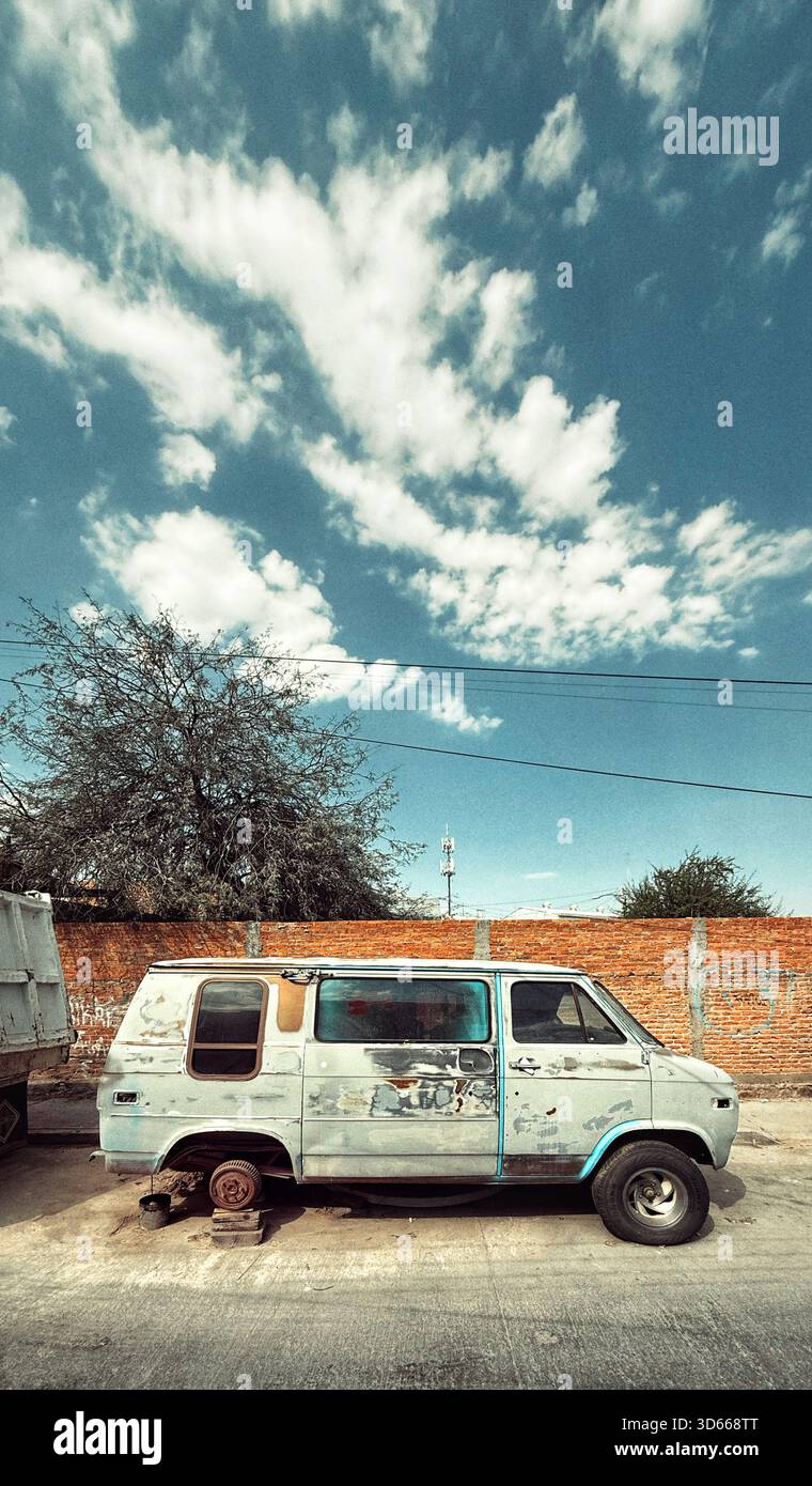 Verlassene und verwitterte Van, fehlende Räder, die auf einer ruhigen Straße unter einem hellblauen Himmel mit verstreuten Wolken geparkt wurden - Smartphone-aufgenommenes Stockfoto