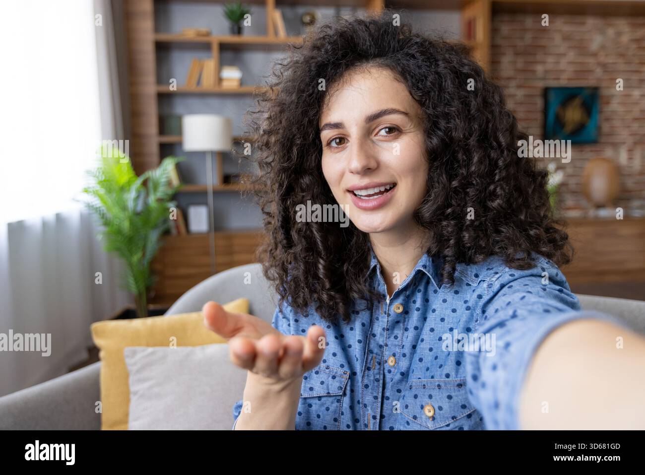Junge Frau mit lockigem Haar, die sich selbst für ein Video filmt oder an einem Videogespräch teilnimmt, lächelt und mit der Hand Gesten bewegt, während sie von einem komfortablen Wohnzimmer aus kommuniziert Stockfoto