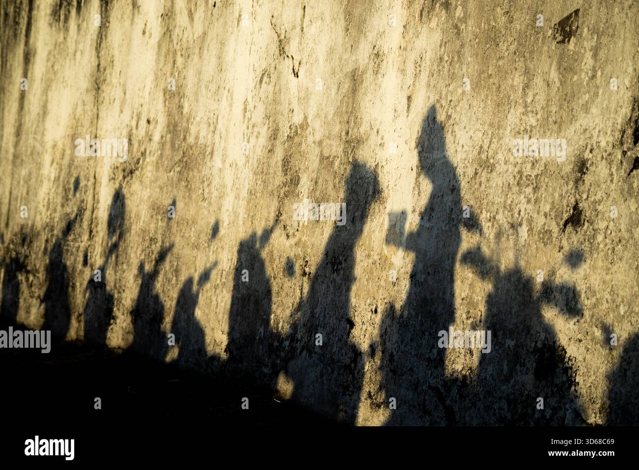 Eine Gruppe von Menschen und Motorrädern wurde auf eine alte, raue, beige-gelbe Wand projiziert. Das intensive, niedrige Nachmittagssonnenlicht erzeugt einen starken Kontrast und d Stockfoto