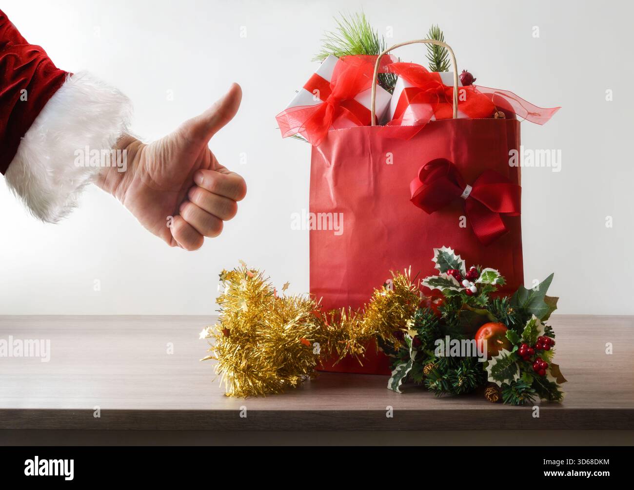 Konzeptionelles Detail des Weihnachtseinkaufs mit einer roten Papiertüte voller Geschenke auf einem Holztisch mit Weihnachtsdekoration und einer Hand, die eine gute Geste macht Stockfoto
