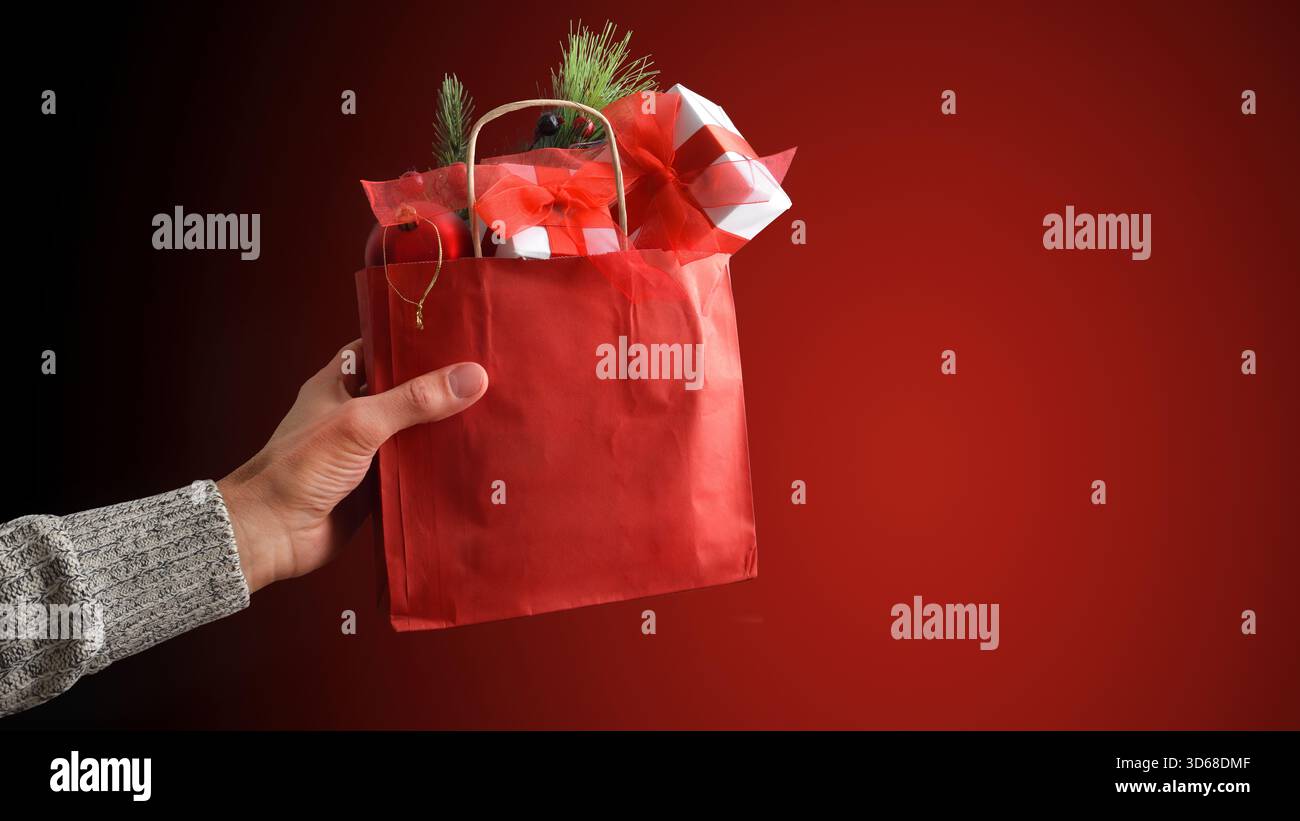 Konzept des Handgebens, bietet eine rote Geschenktüte aus Papier mit Geschenken und Dekorationen mit isoliertem rotem Hintergrund. Vorderansicht. Stockfoto