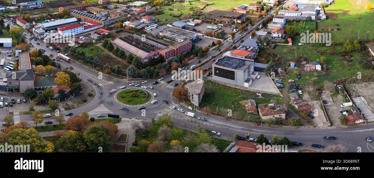 Blick aus der Vogelperspektive auf einen geschäftigen Korçë-Kreisverkehr, umgeben von gemischten Industriegebäuden, Häusern und farbenfrohen saisonalen Bäumen. Stockfoto