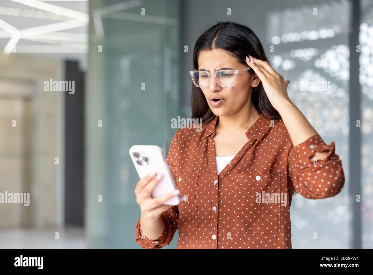 Die junge Frau in einem modernen Büro sieht schockiert und besorgt aus, als sie auf ihrem Smartphone verstörende Nachrichten liest, das Gerät hält und unerwartete schlechte Informationen verarbeitet Stockfoto