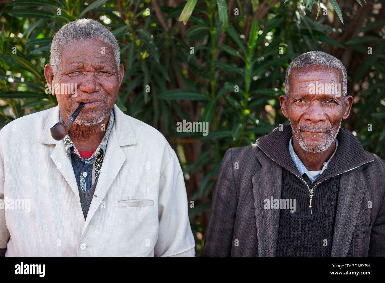 Zwei alte afrikanische Männer stehen draußen im Hof, rauchen Pfeifen, im Dorf an einem sonnigen Tag Stockfoto