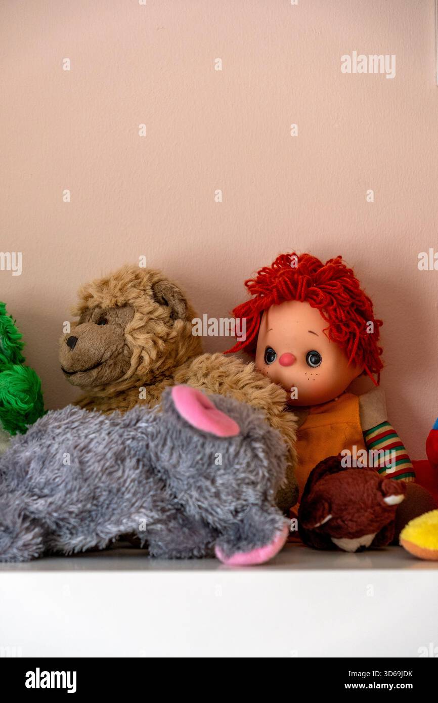 Eine Vielzahl von Vintage-Kuschelspielzeugen säumt ein Regal mit einer rothaarigen Puppe und weichen Tieren in hellen Farben. Die Umgebung ist warm und einladend, perf Stockfoto