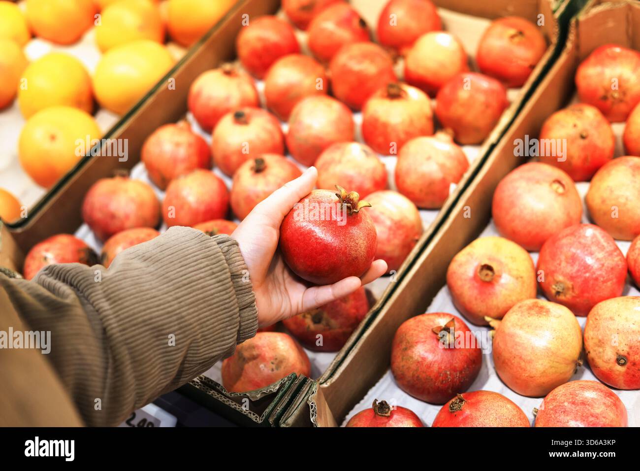 Die Hand einer Person wählt einen Reifen, roten Granatapfel aus einer Vitrine aus, wobei im Hintergrund des Marktes unscharfe Orangen zu sehen sind. Hochwertige Fotos Stockfoto