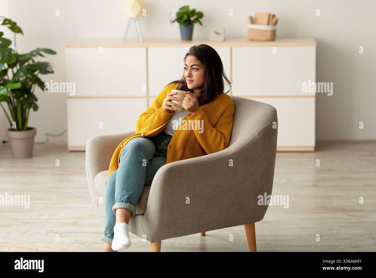 Positive Arab Lady genießt morgens Kaffee, während sie sich am Wochenende in einem gemütlichen Sessel zu Hause entspannt Stockfoto