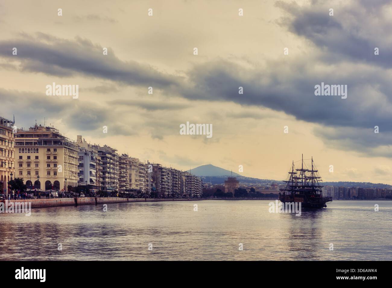 Ein traditionelles Schiff gleitet in Richtung der Küste von Thessaloniki, wo sich der Weiße Turm inmitten moderner Gebäude und dramatischer Wolken erhebt Stockfoto