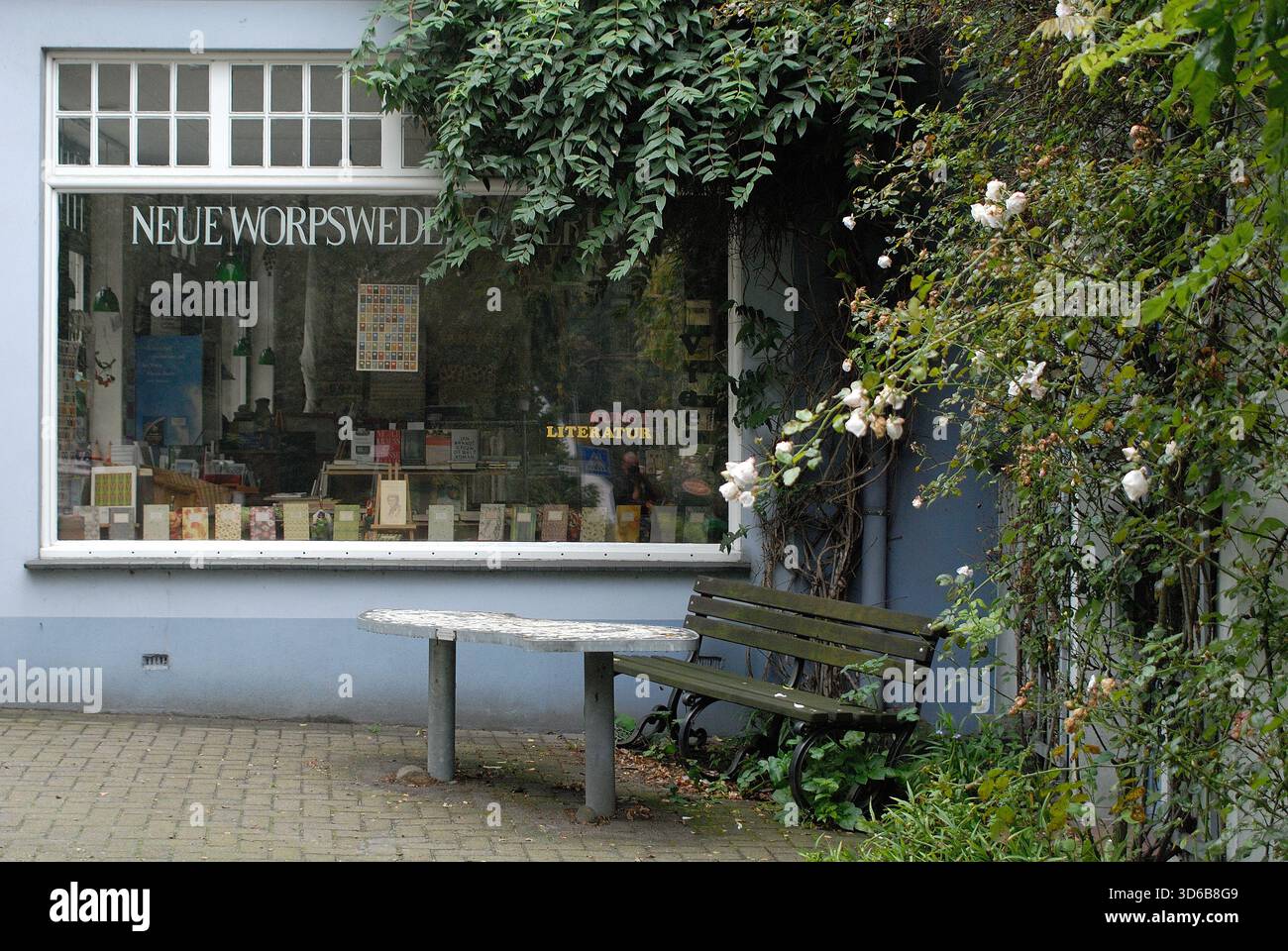 Einladender Außenblick auf die neue Worpsweder Galerie mit einem großen Schaufenster voller Bücher, einer Bank und üppigen Kletterrosen. Stockfoto