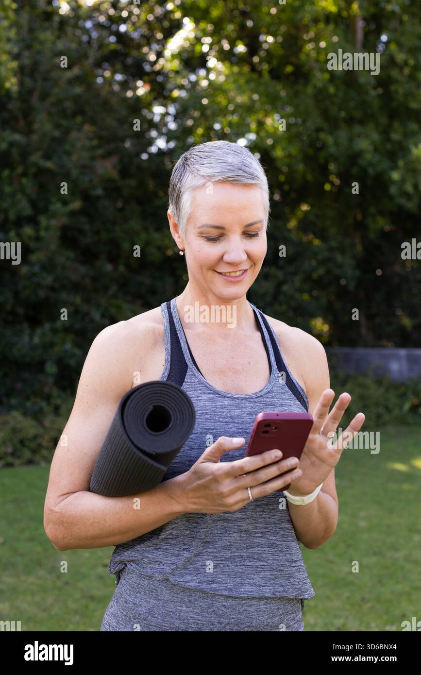 Ältere Frau mit Tanktop, im Hof stehend mit schwarzer Yogamatte, mit rosa Smartphone Stockfoto