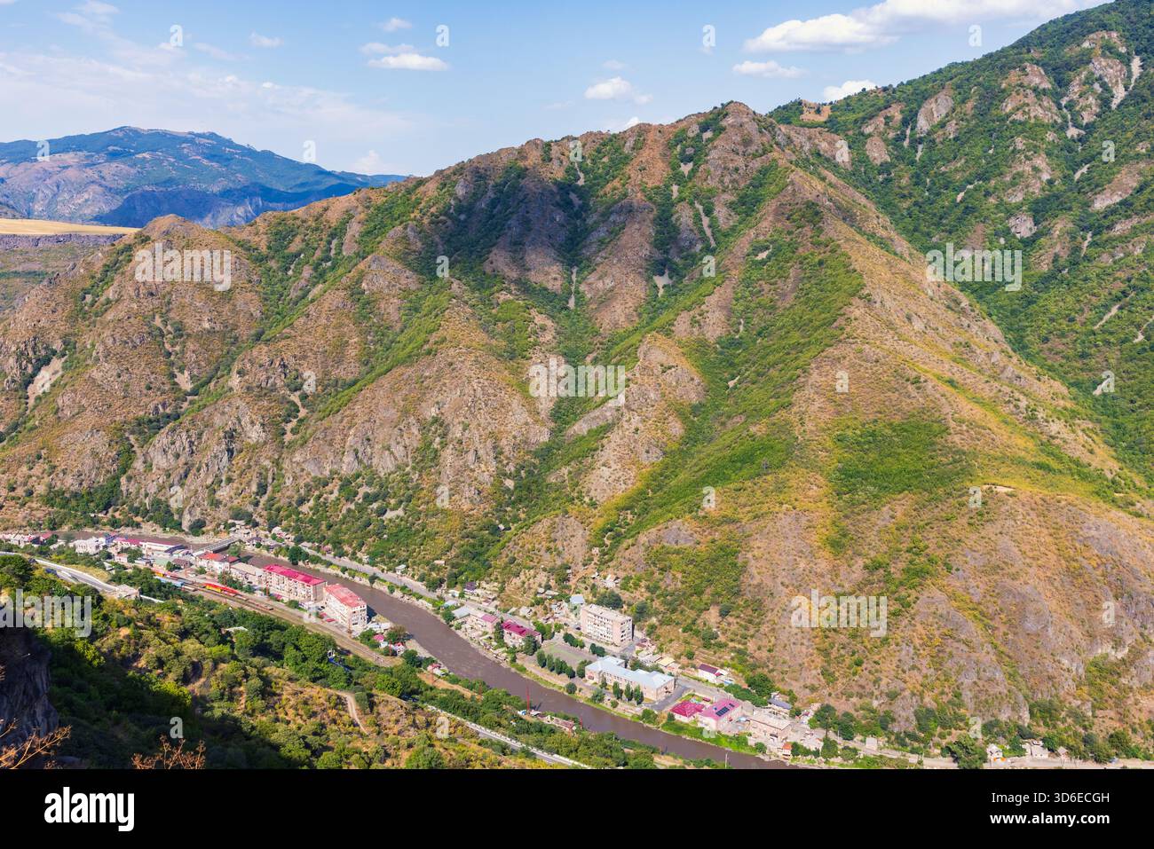 Eine weitläufige Luftlandschaft mit einem Bergdorf, versteckt neben einem gewundenen Fluss, mit zerklüfteten Hängen, grüner Vegetation und klarem blauem Himmel. Al Stockfoto