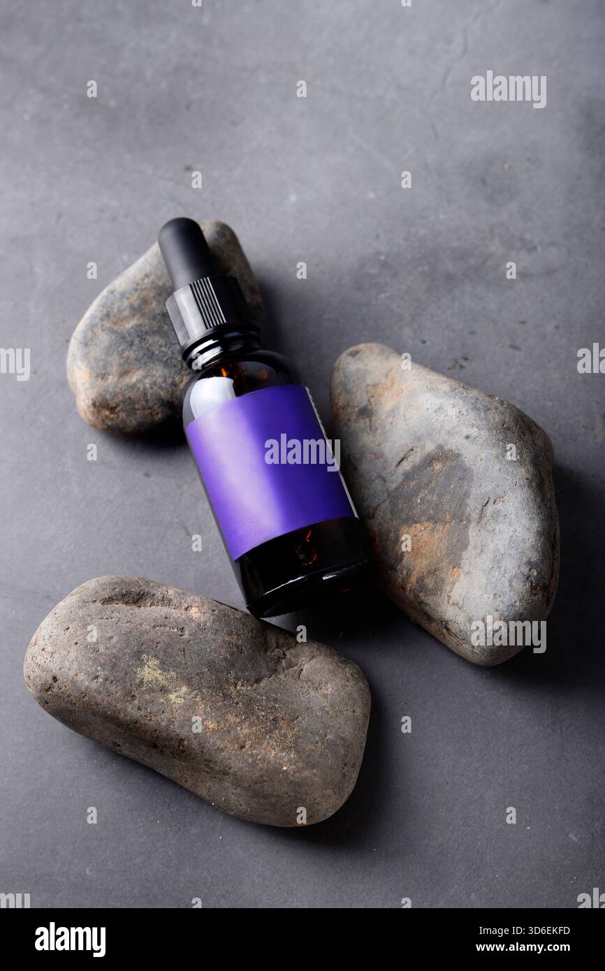 Eine dunkle Flasche ätherischen Öls, gekennzeichnet mit einem violetten Band, ruht zwischen glatten grauen Steinen und symbolisiert natürliche Schönheit und Wellness-Behandlungen Stockfoto
