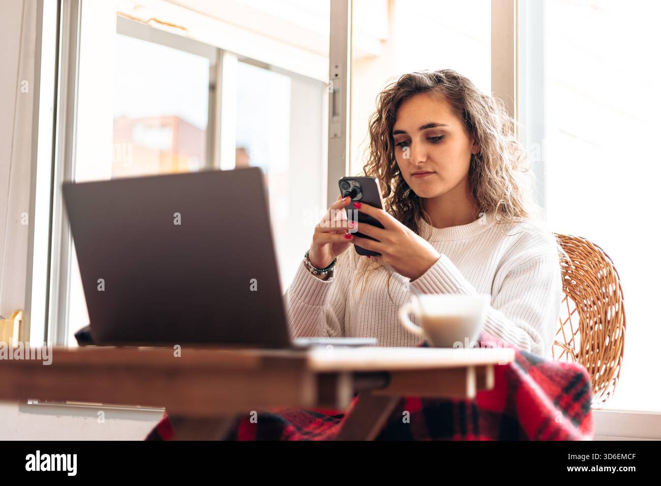 Von unten eine junge Frau in warmem Pullover, die am Tisch mit Laptop sitzt und das Handy surft, während sie von zu Hause aus arbeitet Stockfoto
