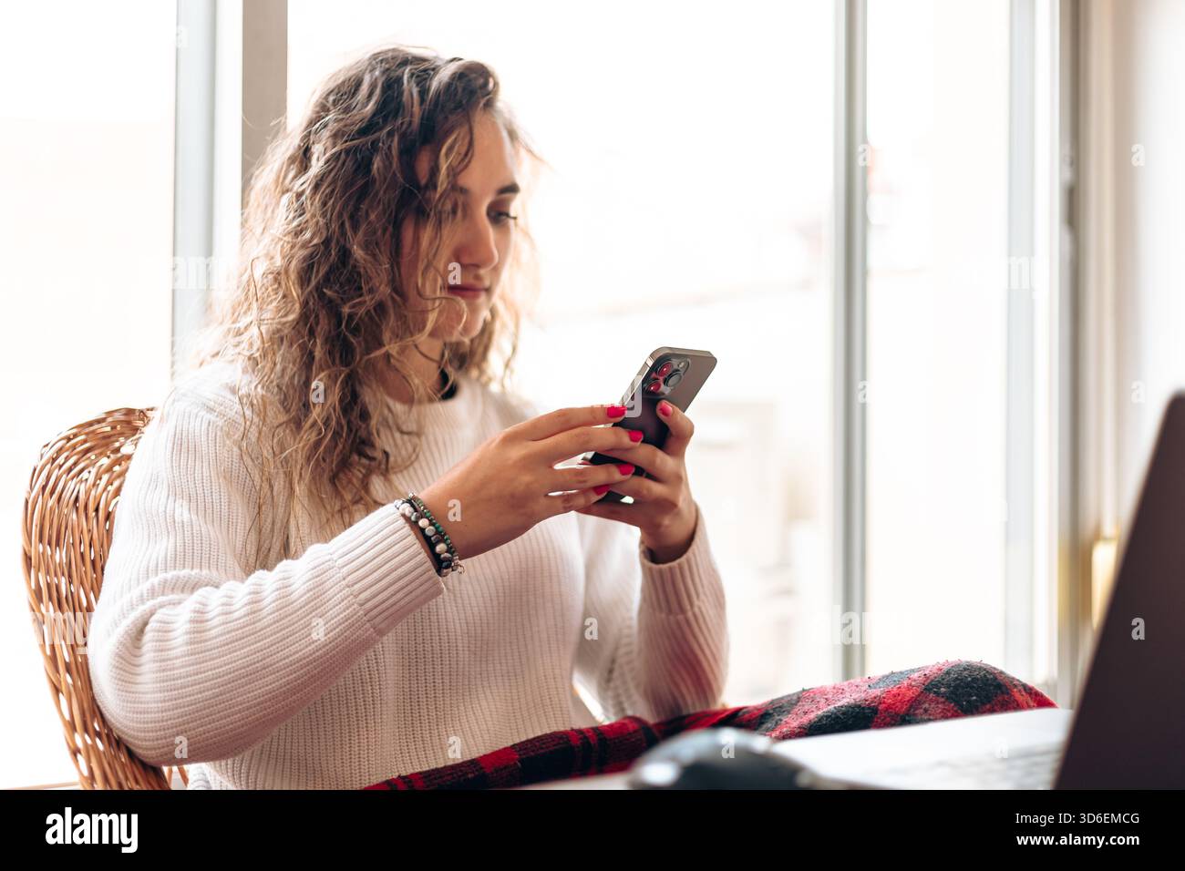 Junge Frau in warmem Pullover, die am Tisch mit Laptop sitzt und das Handy surft, während sie von zu Hause aus arbeitet Stockfoto