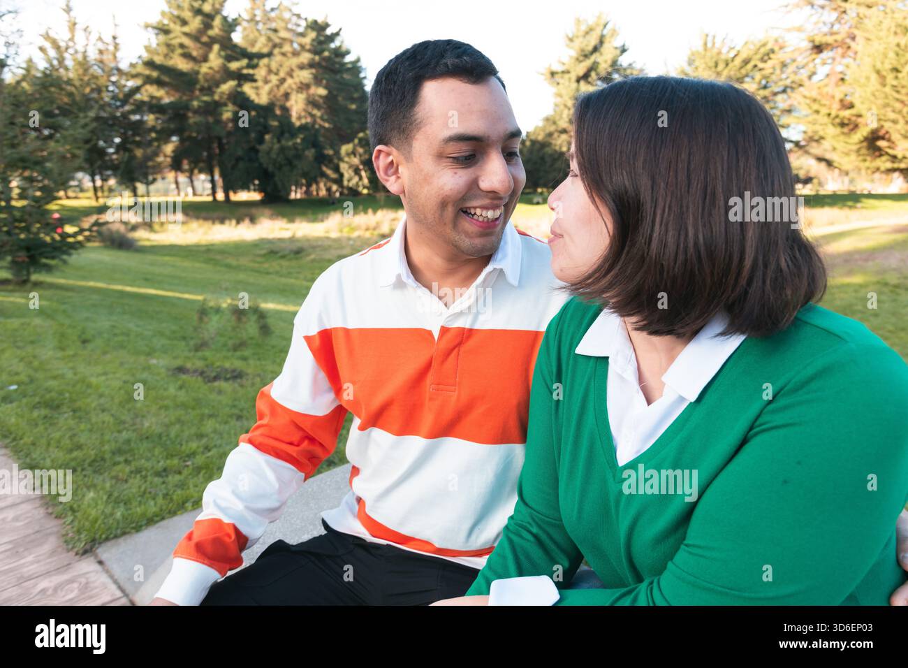 Ein fröhliches, junges lateinisches Paar, das eng zusammen sitzt und sich in einer sonnigen Parklandschaft amüsiert Stockfoto