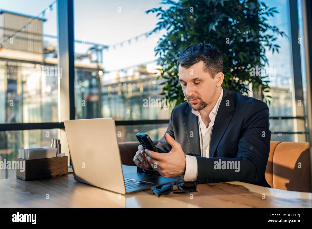 Ein Geschäftsmann im Anzug arbeitet mit einem Laptop an einem Kaffeetisch. Die Umgebung ist entspannt, und das Tageslicht strömt durch große Fenster und schafft so etwas Stockfoto