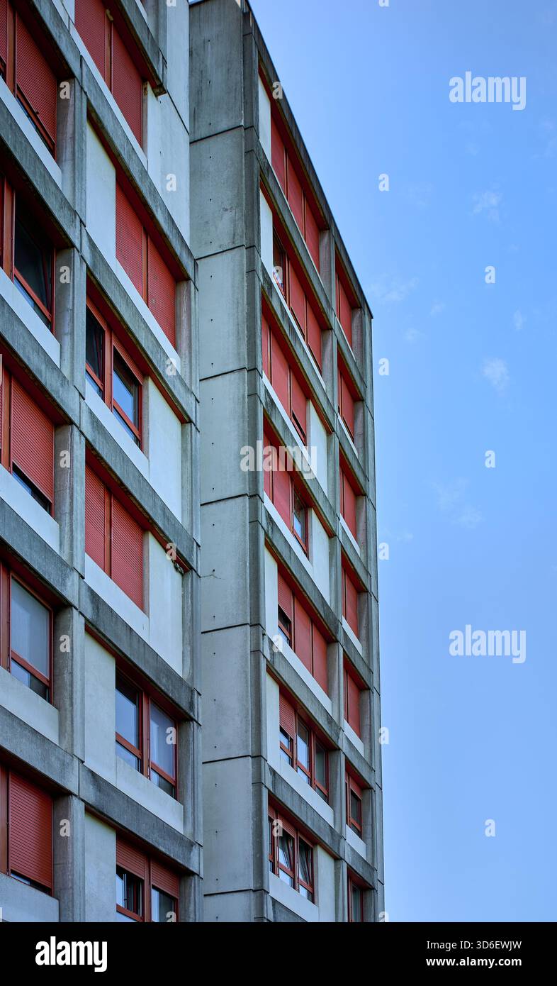Ein brutalistisches Betongebäude mit geometrischen Linien und roten Fensterläden erhebt sich vor einem blauen Himmel und hebt urbane Symmetrie und minimalistisches Design hervor Stockfoto