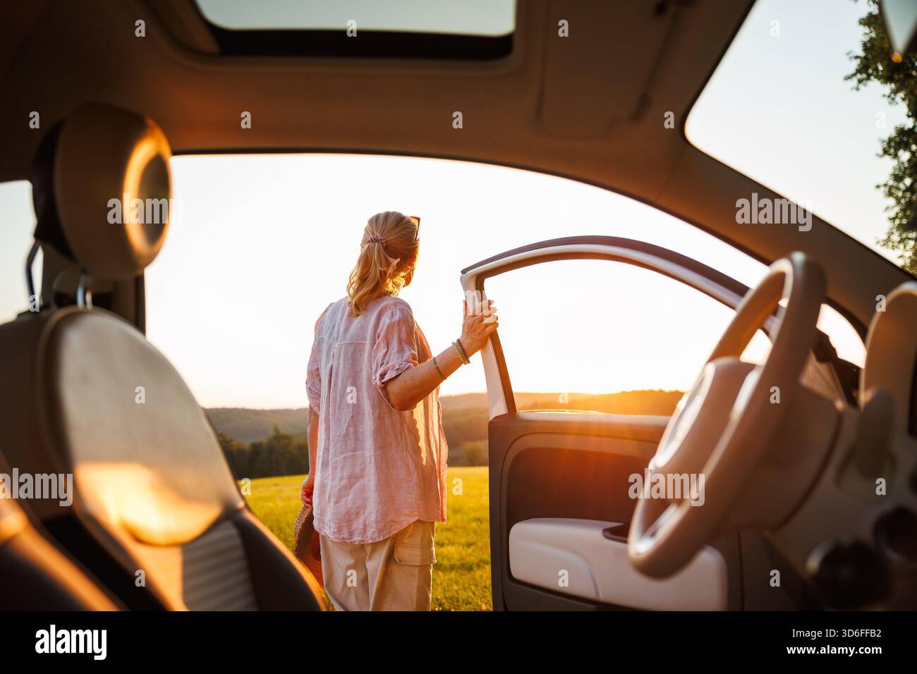 Fröhliche Frau, die aus dem Auto in goldenes Licht des Sonnenuntergangs steigt und das Gefühl von unbeschwertem Reisen während eines Roadtrips im Sommer festhält. Blick vom Innenraum des Fahrzeugs Stockfoto