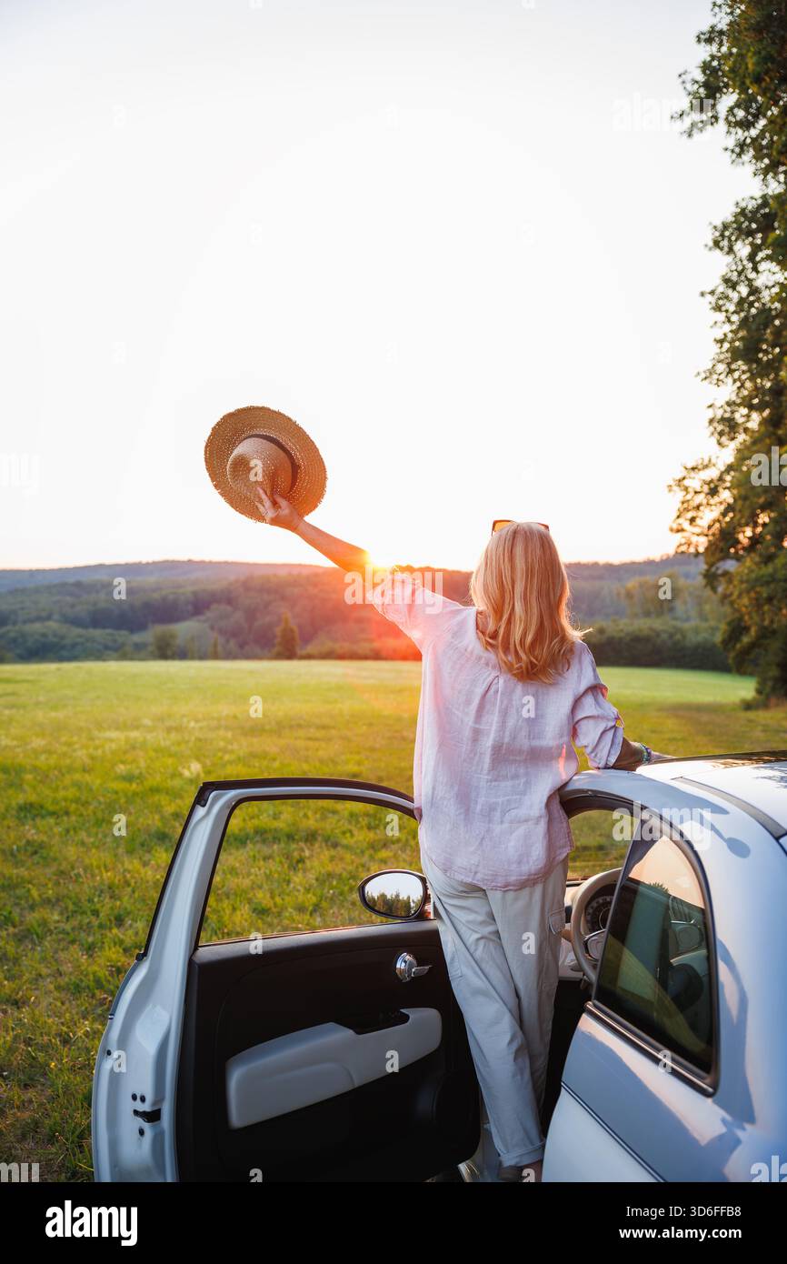 Glückliche Frau, die bei Sonnenuntergang einen Hut in der Nähe des Autos hält, einen Roadtrip im Sommer feiert und Freude am Alleinreiseleben hat Stockfoto