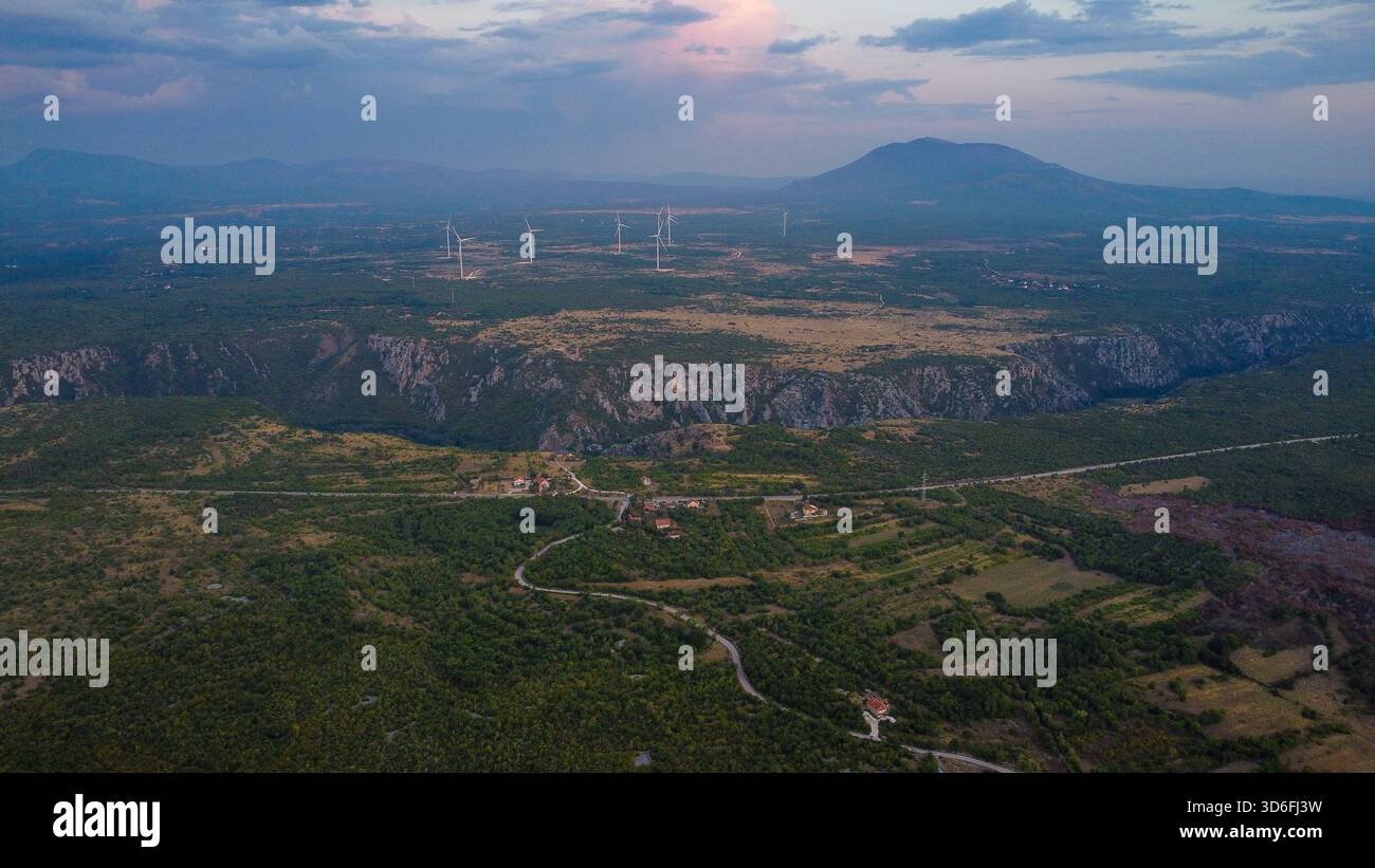 Riesige europäische Landschaft mit modernen Windturbinen und einer steilen Flussschlucht, mit sanftem Gelände und fernen Bergen unter einem dramatischen Abenddämmerhimmel Stockfoto