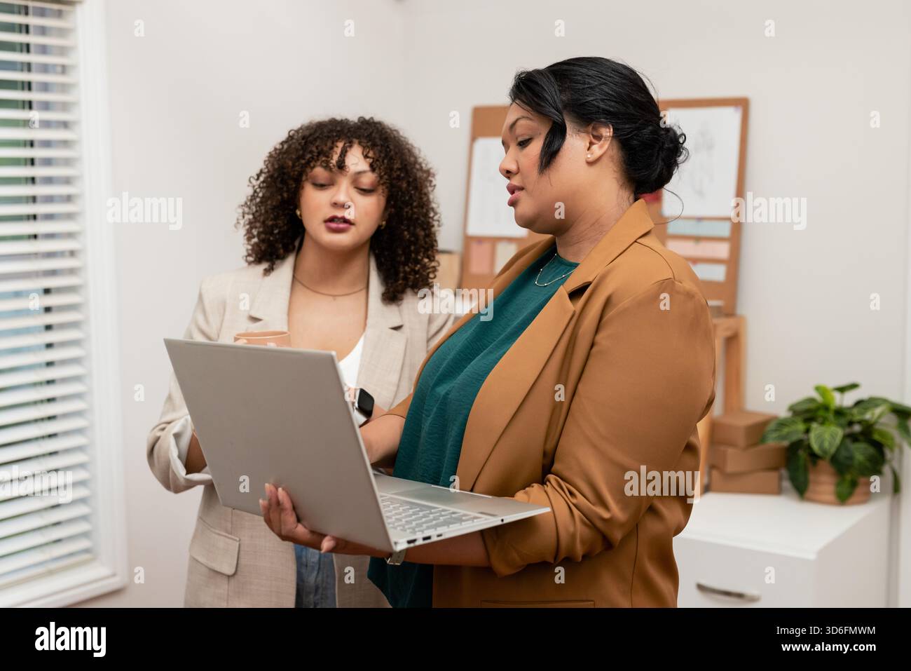 Verschiedene weibliche Kollegen, die im Büro stehen und silberne Laptops und Smartphones mit Korkbrettern untersuchen Stockfoto