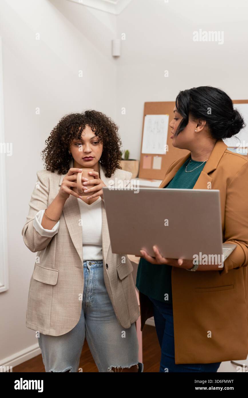 Verschiedene weibliche Kollegen, die Blazer tragen, die einen Becher halten und einen Laptop im kompakten Büro präsentieren Stockfoto