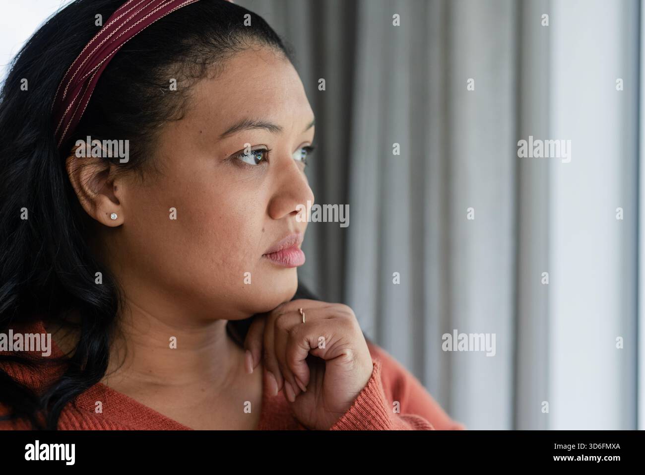 Asiatische afroamerikanische mittelErwachsene Frau, die am Fenster sitzt und Rostpullover und gestreiftes Stirnband trägt Stockfoto